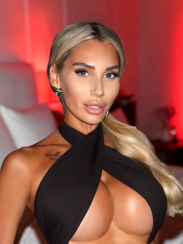 A woman with long blonde hair and striking makeup wearing a black halter top, posing confidently in a warmly lit room with a bed in the background.