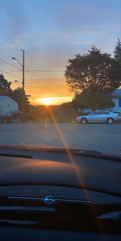 Sunset view from inside a car, featuring a street with trees, houses, a white car, and power lines under a colorful sky.