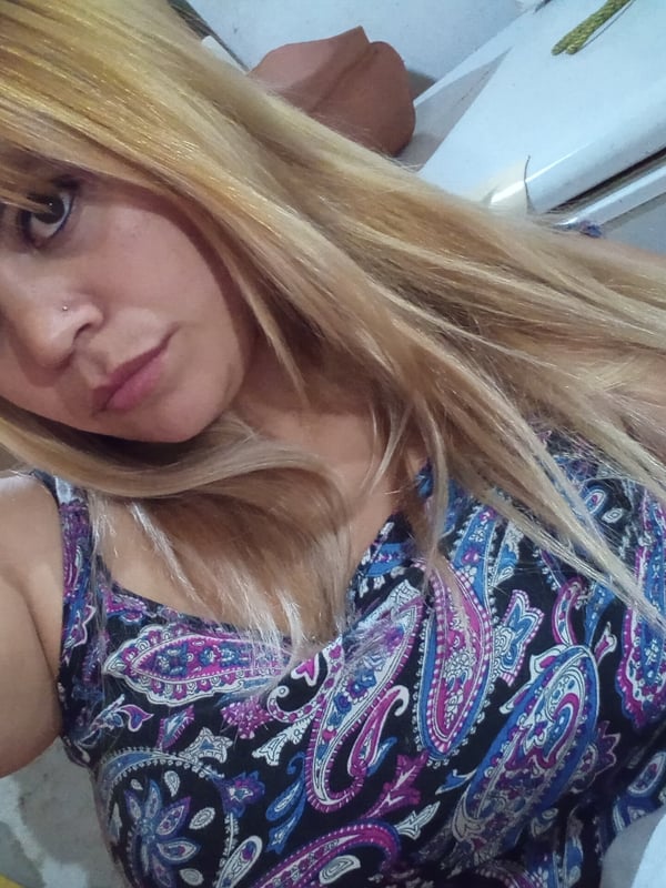 Young woman with long blonde hair and a neutral expression, wearing a colorful strap top, posing indoors near a window.