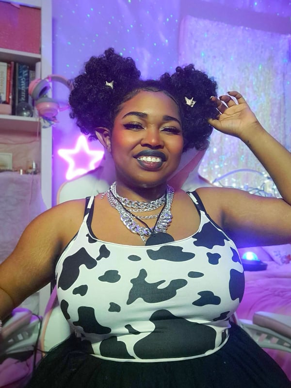 A young woman with curly pigtails smiling in a colorful bedroom with neon lights and playful decor, wearing a cow print top and necklace.