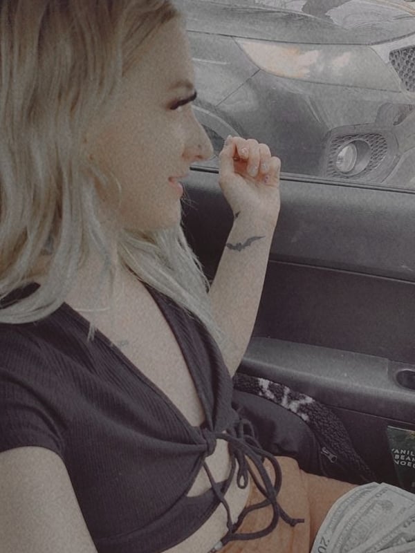 A young woman with blonde hair sits in a car, smiling while holding cash, dressed in a dark laced top and light pants.