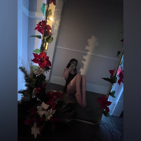 A woman sitting on the floor in a dimly lit room surrounded by holiday plants and decorations, holding a drink.