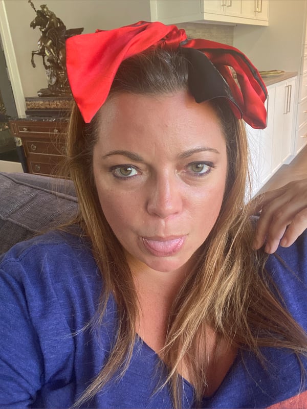 A woman with long brown hair wearing a large red bow headband and a blue top, sitting indoors with decorative background elements.