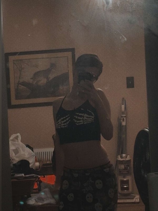 Young woman taking a mirror selfie in a dimly lit room, wearing a black skeleton crop top and patterned pants.