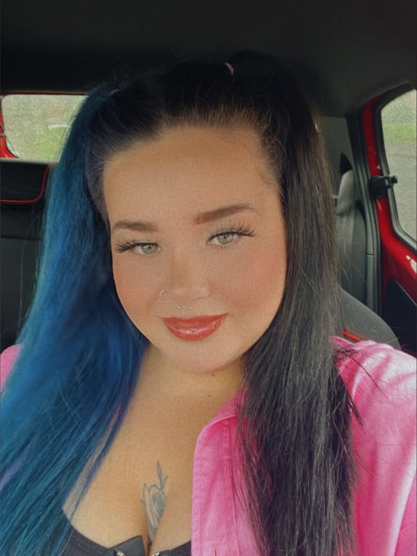 Young woman with long dark hair and blue highlights, wearing a pink zip-up top, posing in a car interior.
