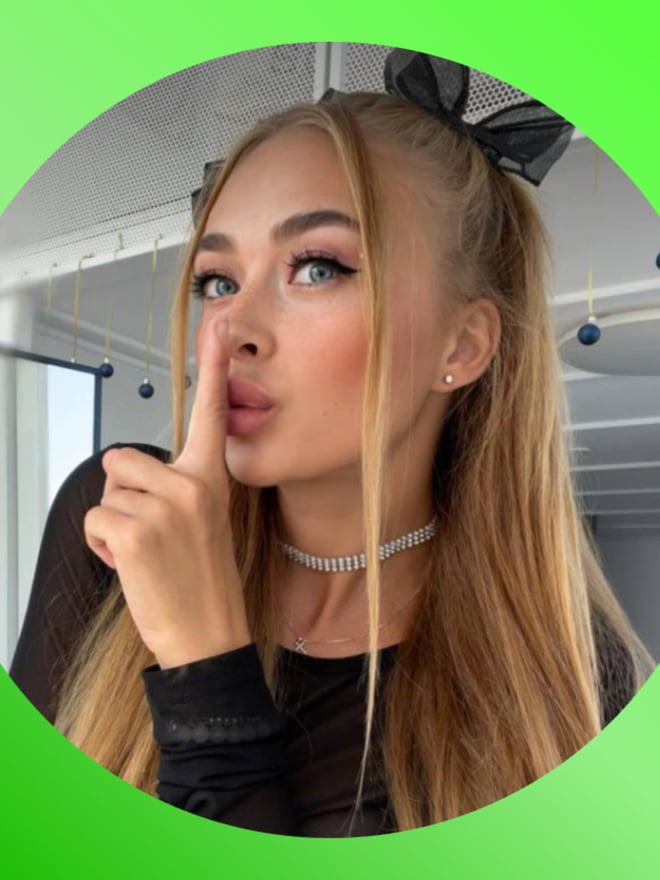 Close-up of young woman with long blonde hair and black bow, making a shushing gesture with finger to lips, wearing a choker necklace.