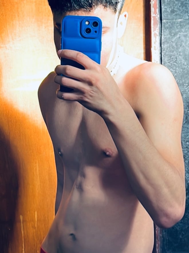 A young man takes a mirror selfie shirtless in underwear, holding his phone in a room with wooden door.