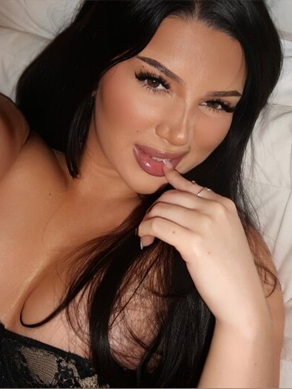 A woman taking a selfie on a bed, wearing lace lingerie and making a playful expression.