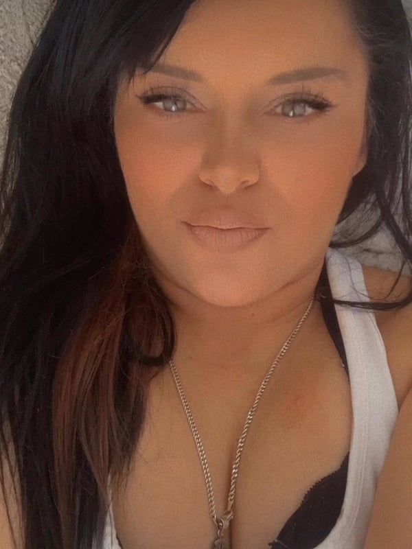 A woman with long dark hair and green eyes smiling at the camera, wearing a white tank top and necklace.