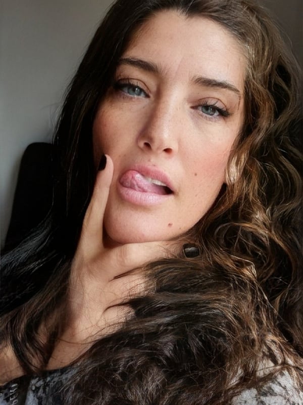A woman with wavy brunette hair and blue eyes, posing with her finger on her lips in a close-up portrait.