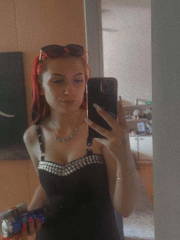 A young woman with red hair and sunglasses on her head, taking a selfie in a black top inside a room with beige walls and a mirror.