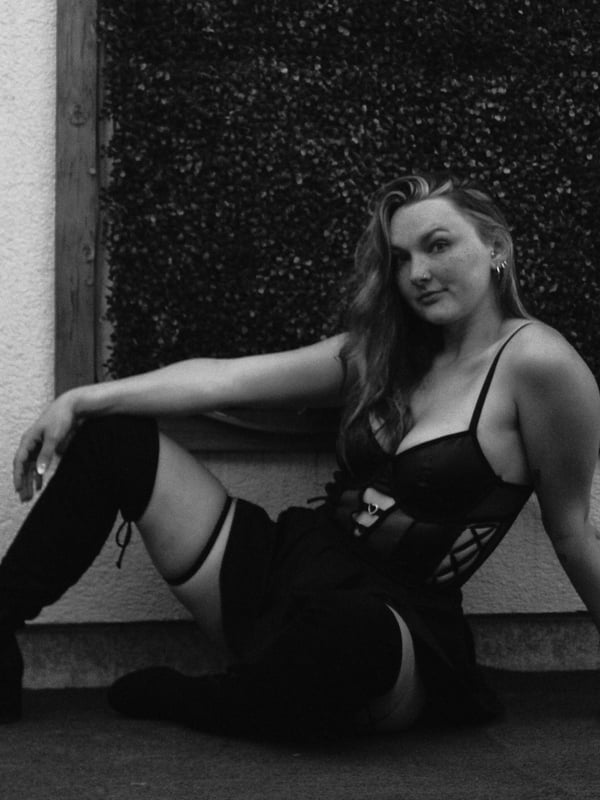 Woman with long wavy hair sitting on the floor in lingerie and thigh-high stockings against a window with greenery.