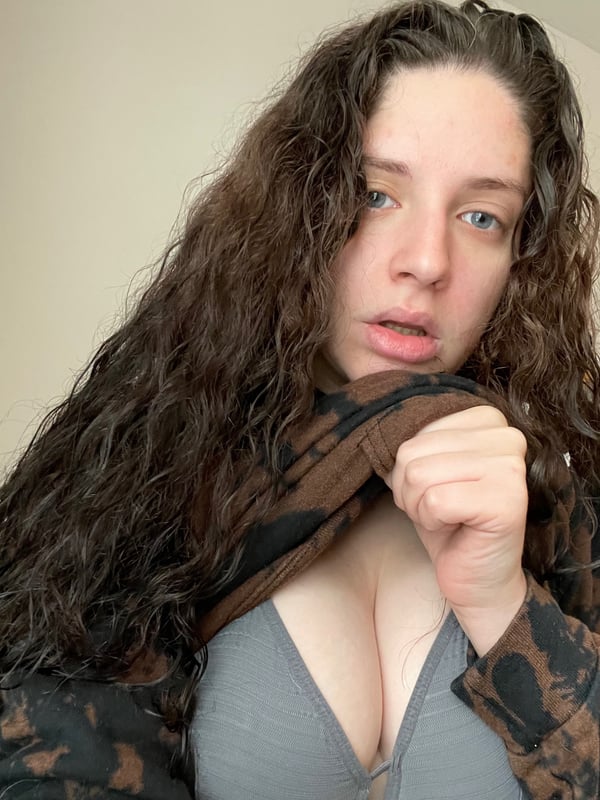 A young woman with wavy dark hair wearing a tie-dye hoodie and gray bra, posing playfully indoors.