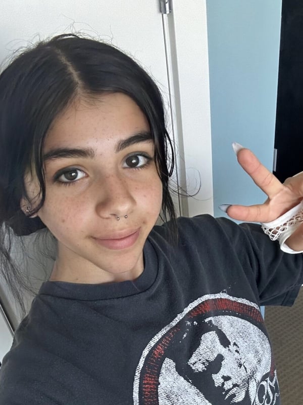 A young woman with dark hair in a ponytail posing with a peace sign hand gesture, wearing a black graphic t-shirt and lace sleeves, standing in a room with white walls and doors.