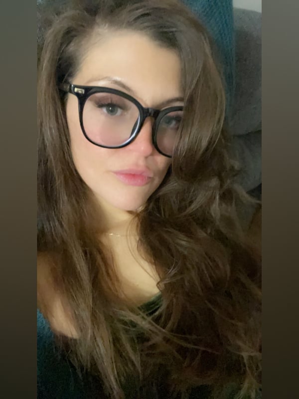 Woman with long brunette hair wearing large black-framed glasses, smiling softly against a teal background.