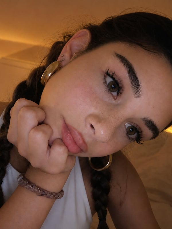 Young woman taking a mirror selfie in a bedroom with string lights, wearing a heart-patterned tank top and red underwear, with long dark hair in braids.