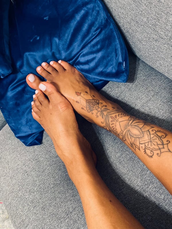 Close-up of bare feet with white-painted toenails positioned on blue pillows against a gray couch background, featuring intricate tattoos on the ankles.