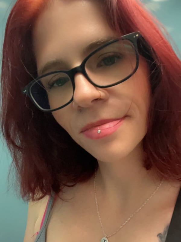 A woman with red hair and glasses smiling at the camera, wearing a tank top in an indoor setting with soft lighting.