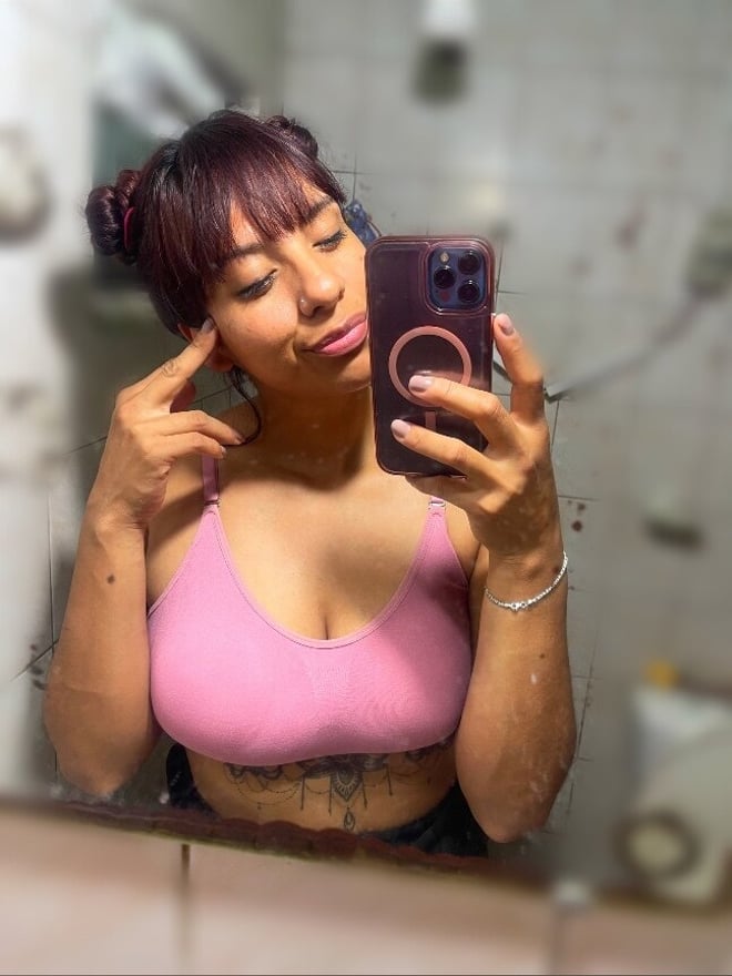 A woman with hair in buns posing in a pink top in front of a bathroom mirror, making a playful gesture while holding a phone.