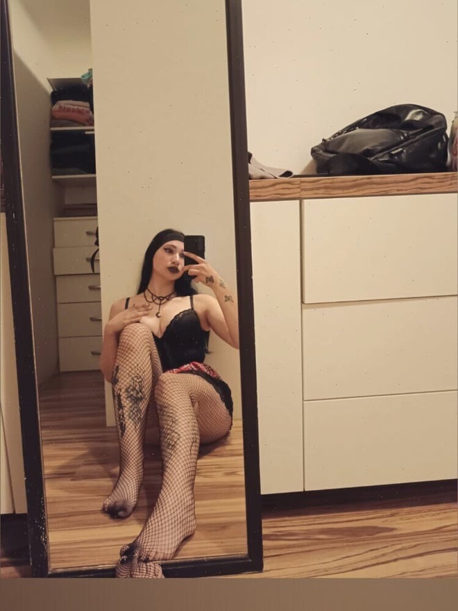 Young woman with long dark hair sitting on the floor in front of a wardrobe mirror, wearing a black top and short skirt, holding her phone.