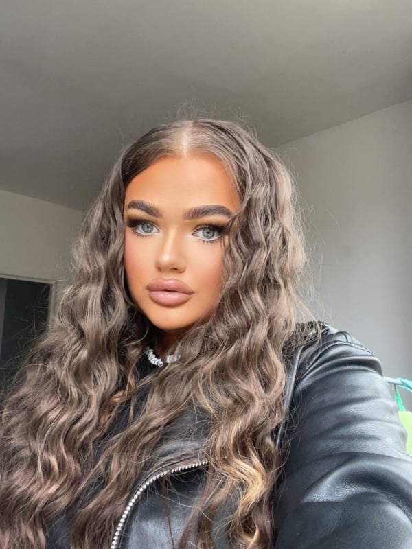 A young woman with long wavy brown hair posing confidently in a black leather jacket against a plain indoor background.