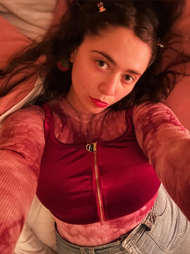 A young woman with brunette hair and red lipstick lying on a bed, wearing a red zip-up top and jeans, smiling at the camera in a close-up selfie.