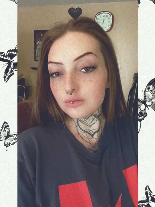 Young woman with brunette hair, freckles, and neck tattoos, posing indoors with a clock in the background, surrounded by a decorative frame of black butterflies.