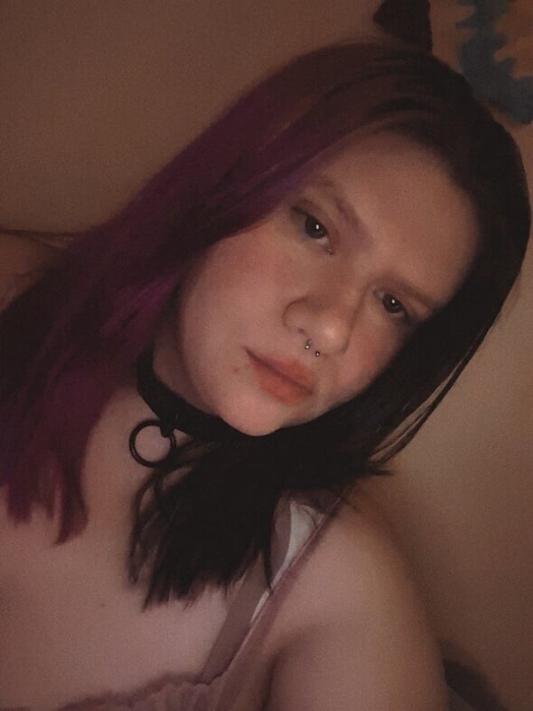 Close-up portrait of a young person with long dyed purple and black hair, wearing a choker necklace, against a dimly lit indoor background.