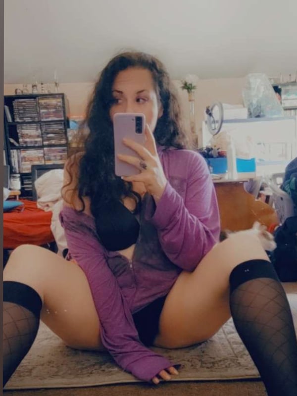 A woman taking a selfie in a purple sweater and black stockings, sitting on the floor in a cozy room.
