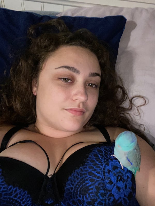 A woman with curly hair lying on a bed in a blue lace bra, holding a small colorful bird on her chest.