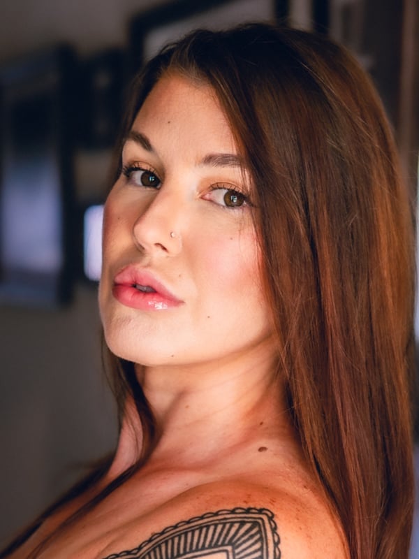 Close-up portrait of a woman with long brown hair, tattoos on her shoulder, and subtle makeup, smiling softly in an indoor setting.