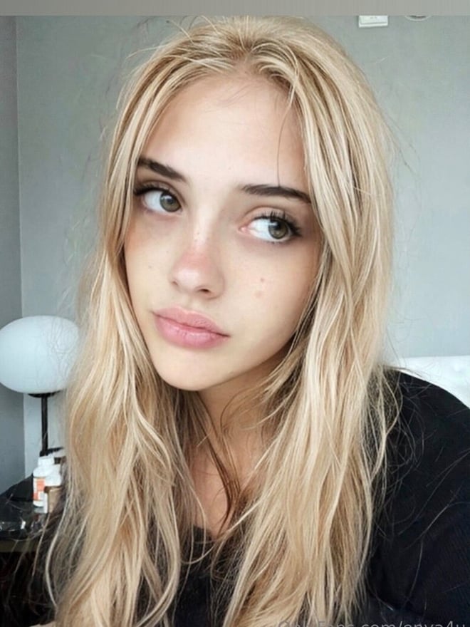 A blonde woman taking a close-up portrait indoors, wearing a dark top.