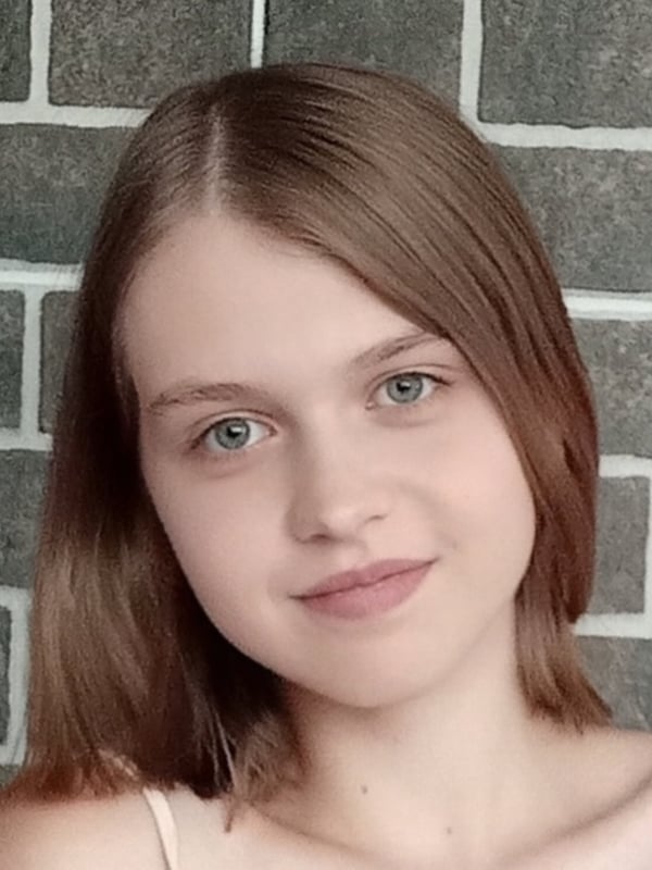 Close-up portrait of a young woman with shoulder-length brown hair and blue eyes, smiling gently while wearing a light-colored top against a brick wall.