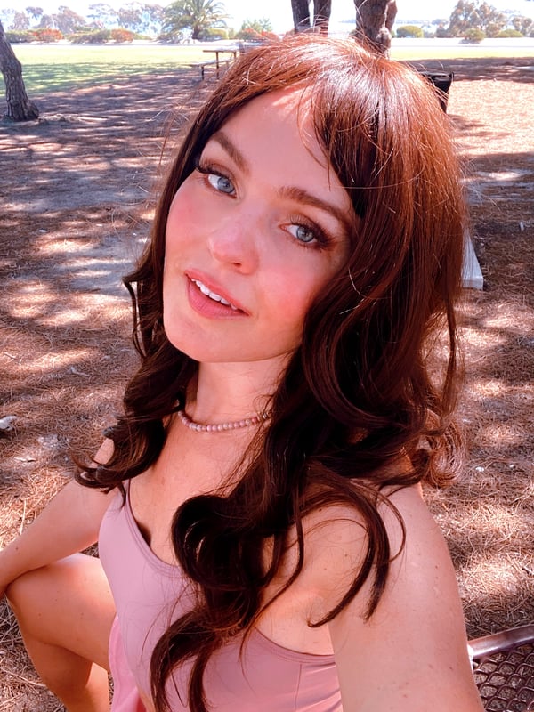 A woman smiling outdoors in a park, wearing a pink top with wavy red hair and natural sunlight.
