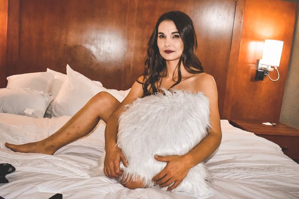 A woman with long dark hair sitting on a bed in a warmly lit bedroom, holding a large white fluffy pillow while posing confidently.