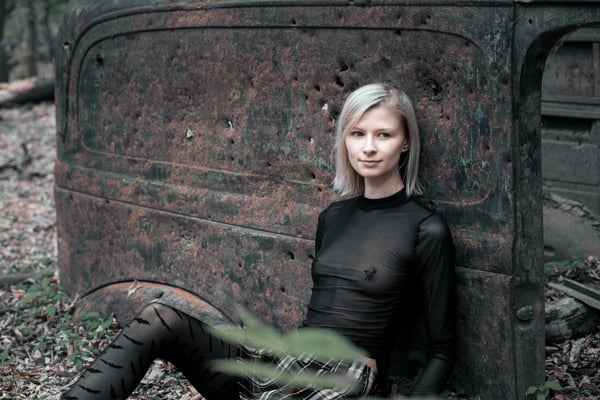 A woman with blonde hair posing in a sheer black outfit and leggings next to an old rusted car in a wooded area.