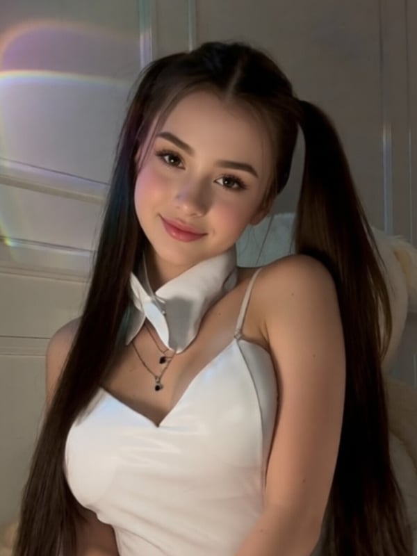 A young woman with long dark hair in pigtails wearing a white top and fishnet stockings, posing indoors.