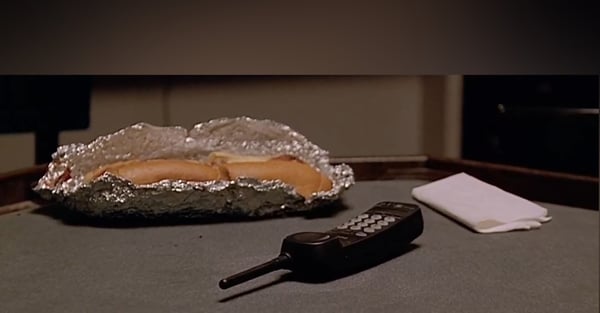 A pair of hot dogs wrapped in aluminum foil placed on a table next to a cordless phone.