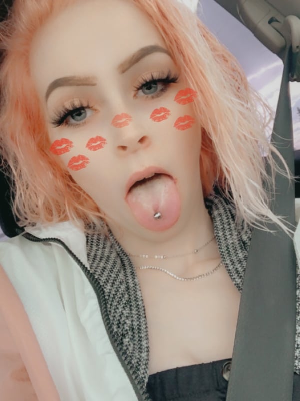 A young woman with pink hair and makeup sitting in a car, wearing a zip-up jacket and showing her tongue with a piercing.