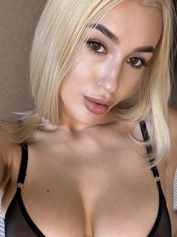 Young woman with blonde bob haircut wearing a sheer black top, posing confidently in a bedroom setting.