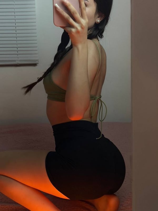 Young woman with long dark hair taking a mirror selfie in a bedroom, wearing a light pink tank top and white bottoms while kneeling on a fluffy pink rug.