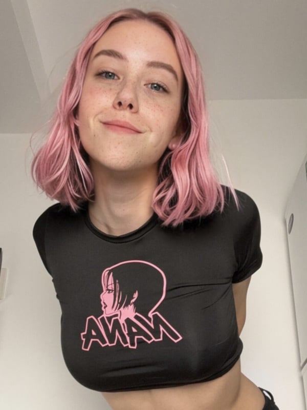 A young person with pink hair wearing a black crop top, standing indoors near a refrigerator.