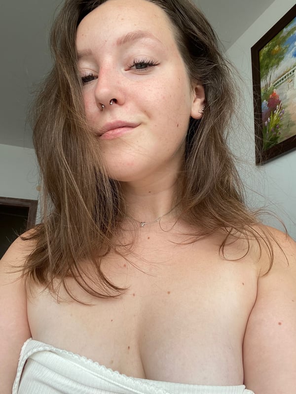 Close-up portrait of a young woman with long brown hair smiling softly while lounging shirtless in a casual indoor setting with artwork on the wall.
