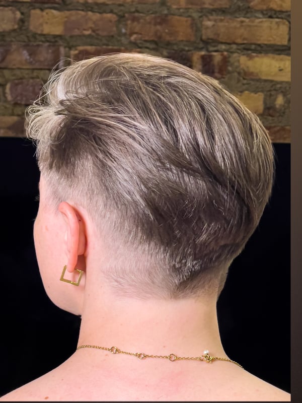 Person with short blonde undercut hairstyle viewed from the back against a brick wall background, wearing gold hoop earrings and a delicate necklace.