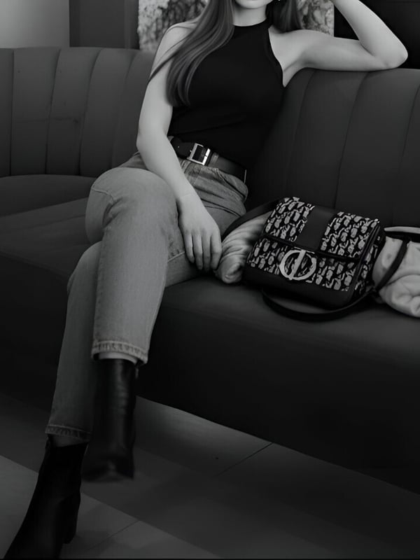 A woman with long hair sitting on a couch in a black halter top, light pants, and boots, holding a designer handbag.
