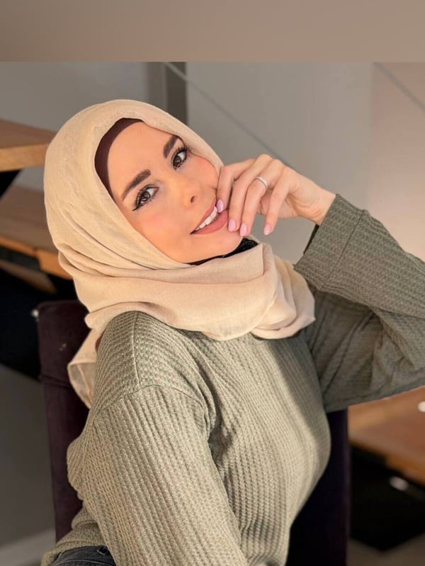 A smiling woman wearing a headscarf and casual clothing poses thoughtfully indoors.