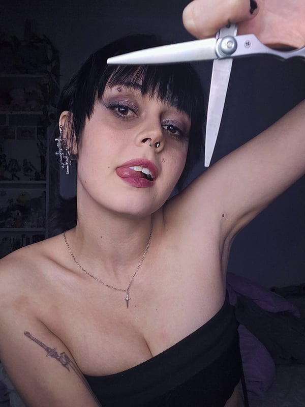 Close-up of a person with short black hair and makeup holding scissors up to their face, wearing a black top, against a bedroom background.