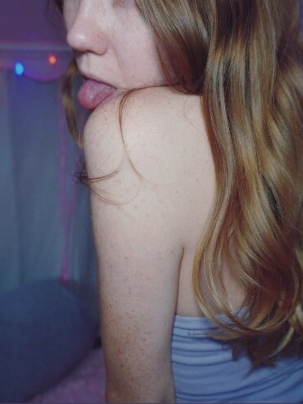 A young woman with freckled skin and wavy reddish hair, sitting inside a pink tent with colorful lights, making a playful expression.