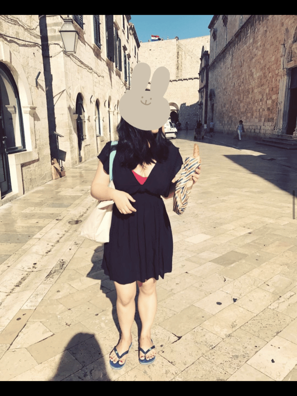 A woman in a black dress holding a banana stands on a cobblestone street in an old European-style town, with a gray bunny sticker covering her face.
