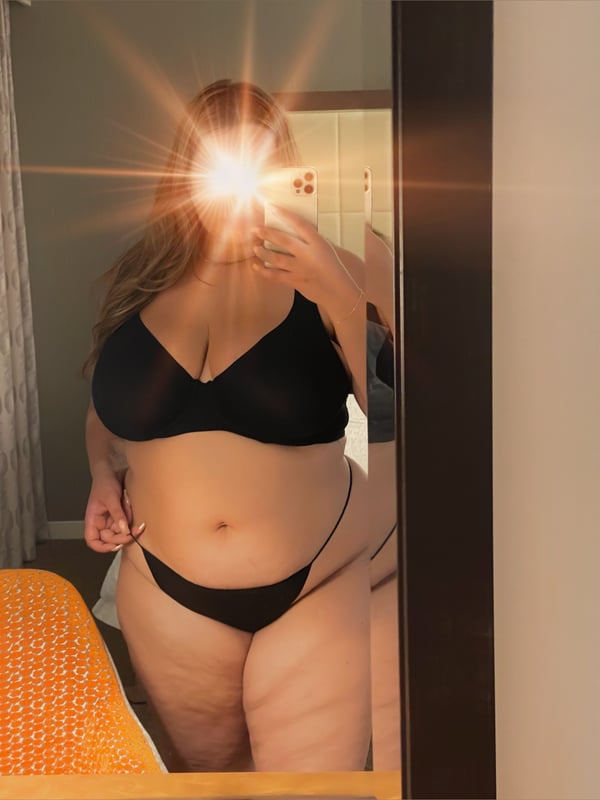A person taking a mirror selfie in a bathroom, wearing black lingerie, with long wavy hair and a glowing filter effect.